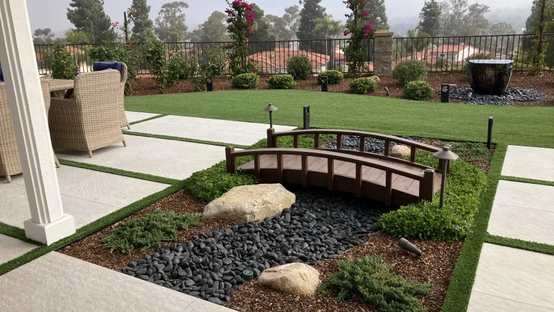 Modern backyard with artificial grass, a small decorative bridge over black pebbles, and outdoor seating area.