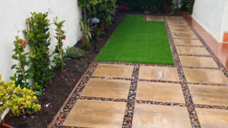 Garden pathway with square stone tiles, planted garden bed, artificial grass strip, and decorative rocks in a backyard.