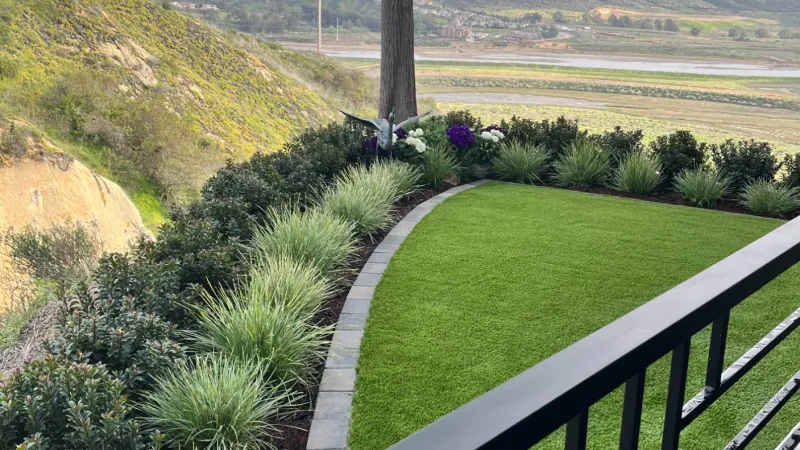 Balcony overlooking landscaped garden with artificial turf, ornamental grasses, a tree, and distant valley views under a cloudy sky.
