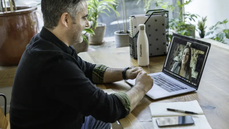 Man video conferencing with woman on laptop at wooden desk with notebook, phone, and water bottle