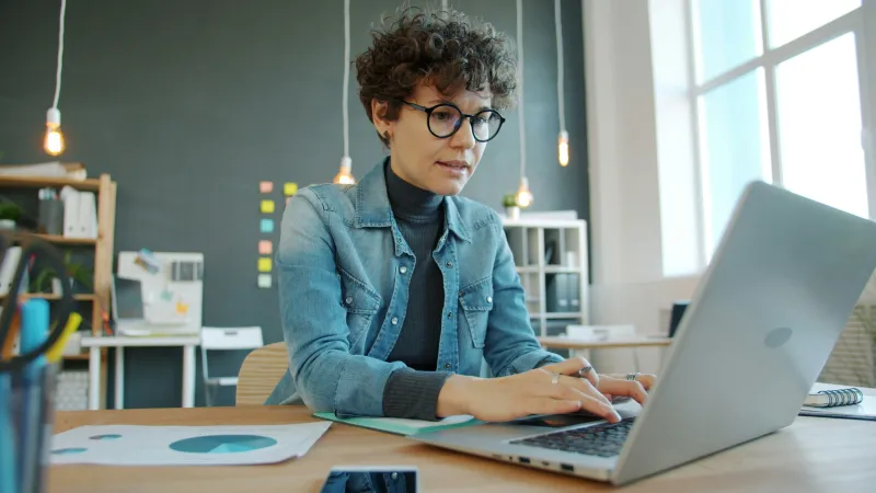 Young woman wearing glasses working on laptop at desk with documents in modern office space with large windows.