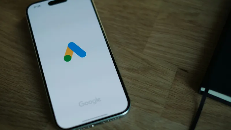 Smartphone displaying Google Ads logo on screen on a wooden table with a notebook.