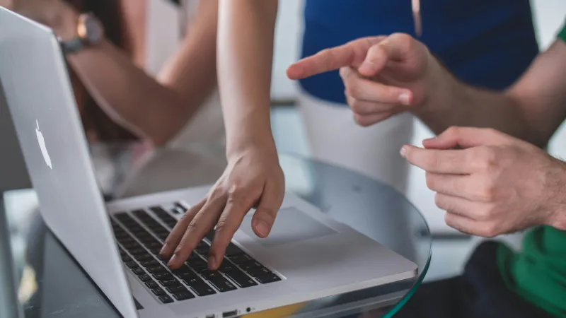 Hands of people collaborating and pointing on a laptop keyboard during a meeting at a glass table.