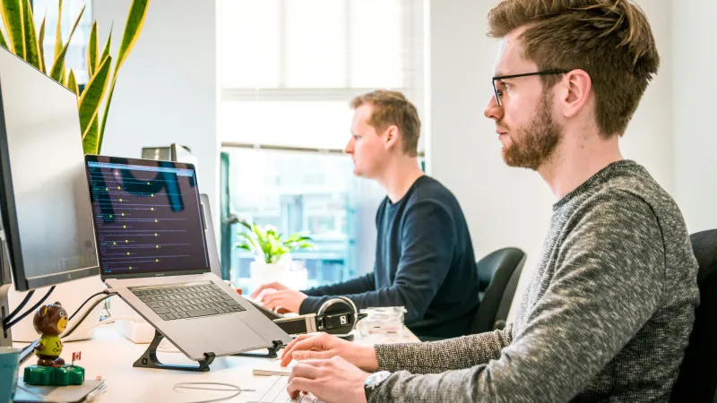 Two men working at computers in a bright office with plants and modern tech setup.