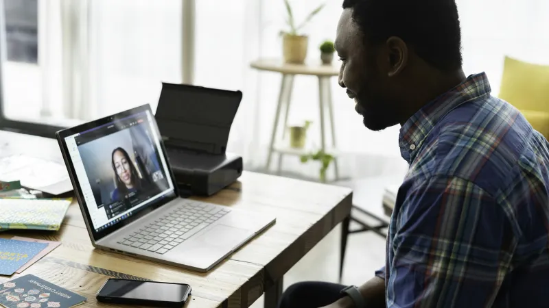 Man in plaid shirt video chatting with woman on laptop in bright home office setting.