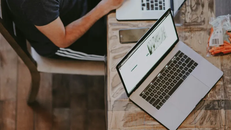 Two people working on laptops side by side at a wooden table with headphones and smartphone nearby