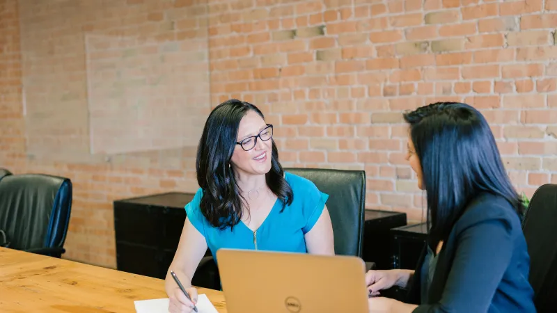 Two professional women collaborating at a wooden conference table with laptop and notebook in modern office.