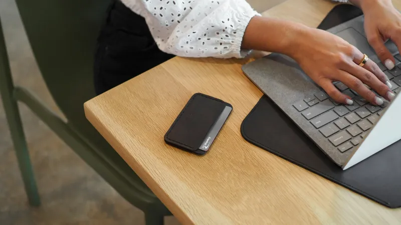 Person typing on laptop at wooden table with black wallet placed nearby in modern workspace