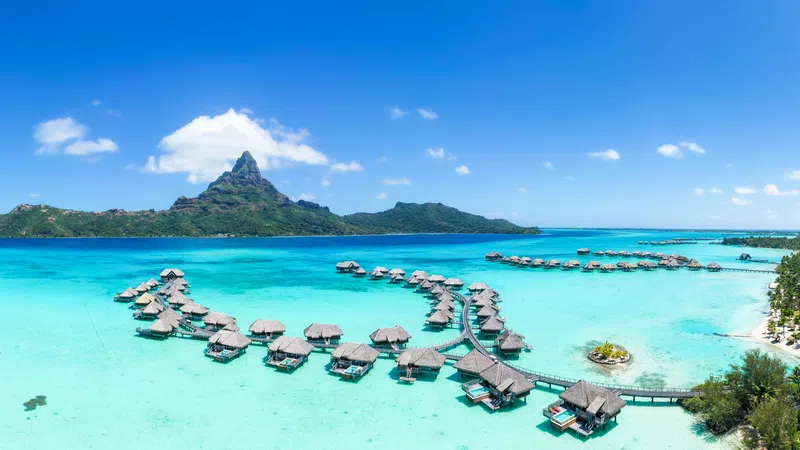 Aerial view of overwater bungalows with turquoise lagoon, tropical island lush greenery, and mountain under blue sky.