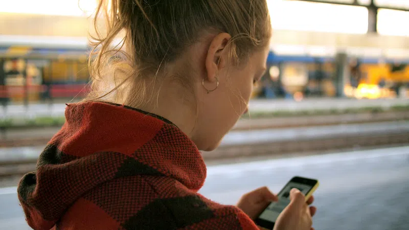 Woman in red plaid jacket using smartphone at train station platform during daytime.