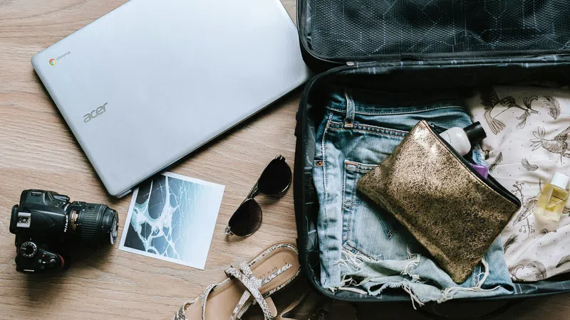 Open suitcase packed with denim shorts, toiletry bag, perfume bottle, next to laptop, camera, sunglasses, and sandals on wooden floor