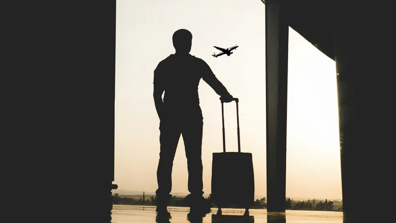 Silhouette of a traveler with suitcase watching an airplane take off through airport window at sunset.