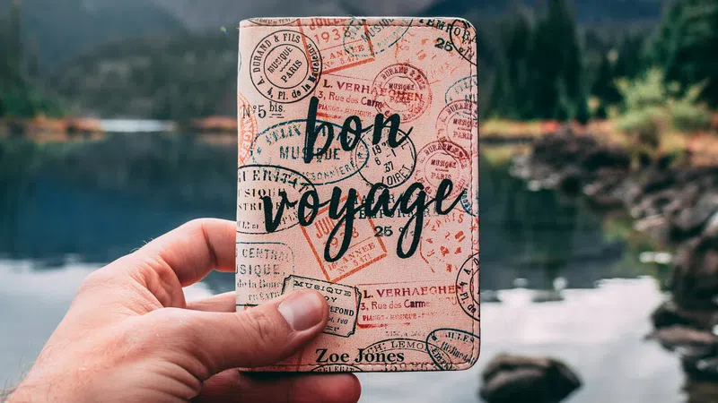 Hand holding a passport cover with bon voyage text against a lake and mountain background on a cloudy day