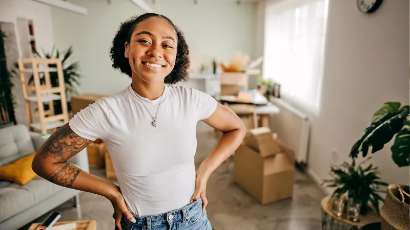Smiling woman with tattoos stands confidently in a bright living room with moving boxes and plants.