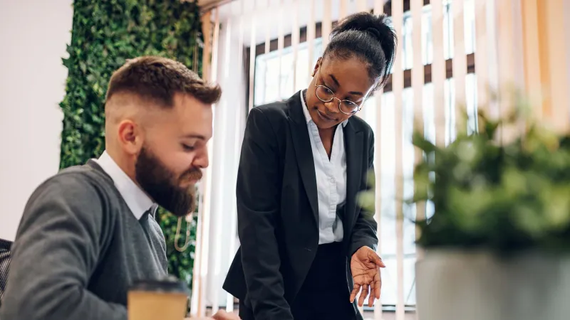 Two colleagues discussing and reviewing documents in a bright modern office with plants and coffee cup.