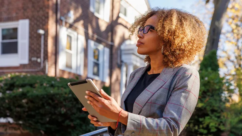 Confident woman with curly hair and glasses using a tablet outside a brick building on a sunny day