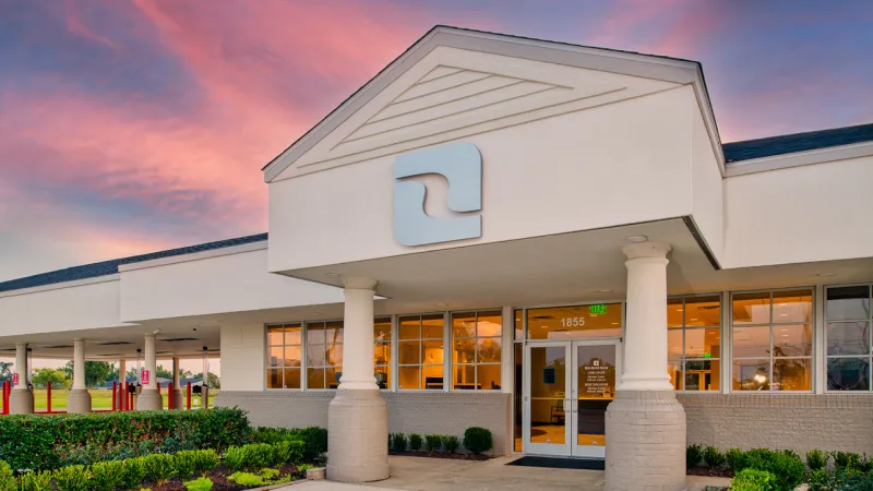 Modern white building entrance with pillars and a logo under a vibrant sunset sky at dusk
