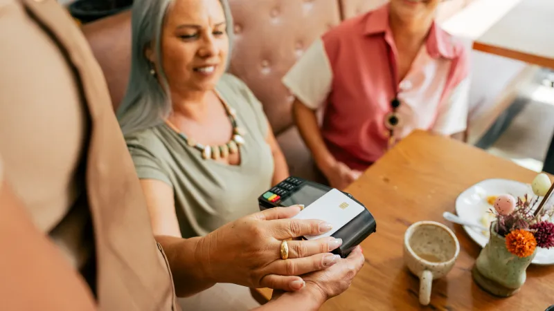Woman paying contactless with card at cafe table while another woman watches and smiles