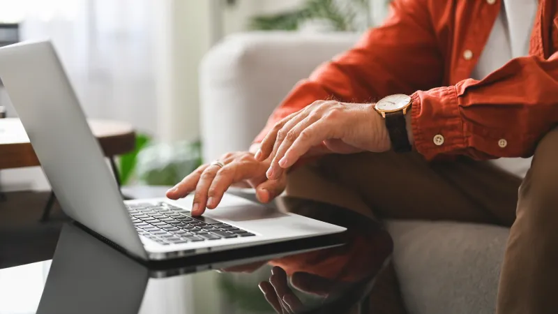 Close-up of a person wearing a watch typing on a laptop keyboard at a reflective glass table indoors.