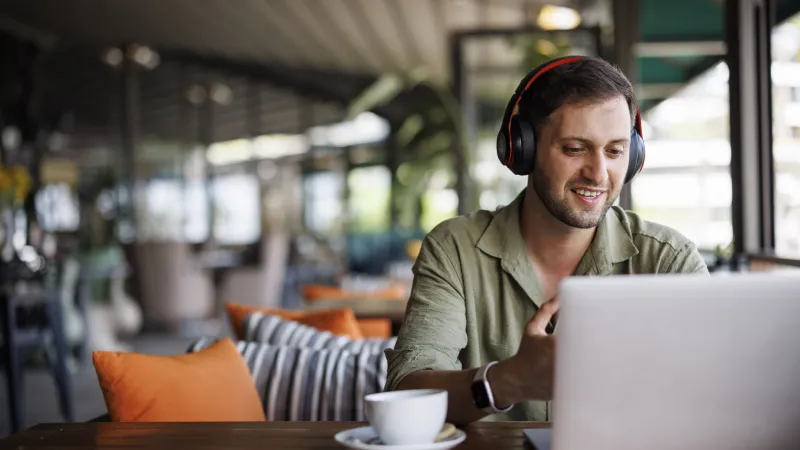 Smiling man wearing headphones using laptop in cozy cafe with coffee cup on table and blurred background