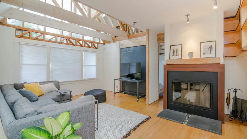 Bright living room with gray sectional sofa, wooden floors, fireplace, TV, and decorative plants under exposed beams