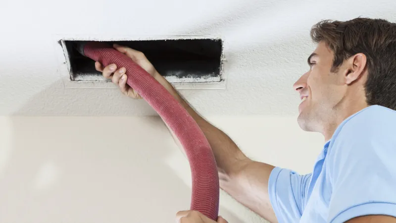 Technician cleaning a ceiling air duct with a vacuum hose, ensuring better air quality.