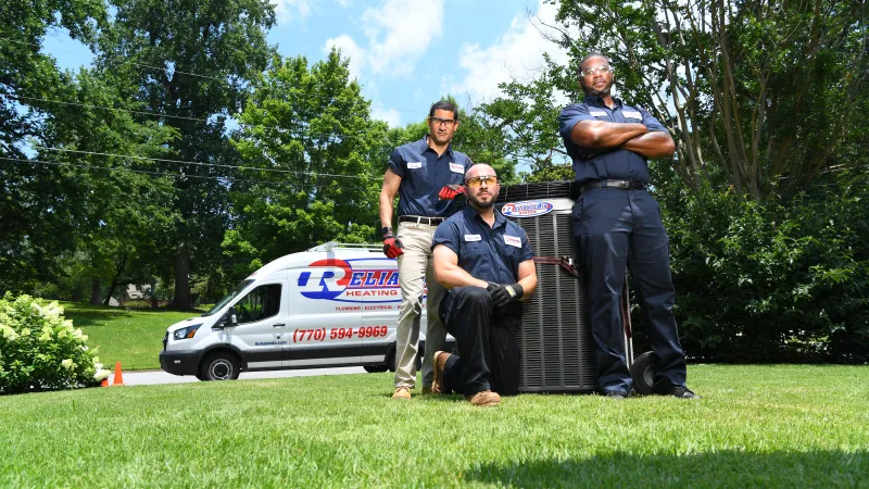 Three HVAC technicians posing with a heating unit on green lawn in front of service van on a sunny day.