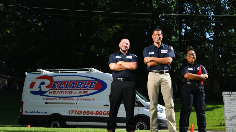 Three smiling HVAC technicians standing confidently in front of a Reliable Heating & Air service van.