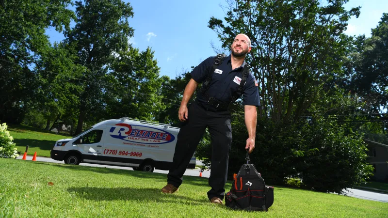 HVAC technician standing on grass with tools, Reliable Heating & Air van in the background under clear sky.