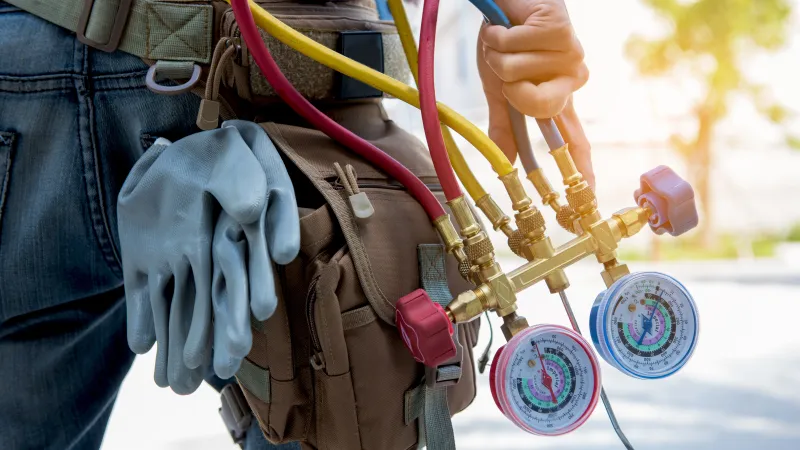 HVAC technician holding manifold gauge set with colored hoses and gloves hanging from tool belt.