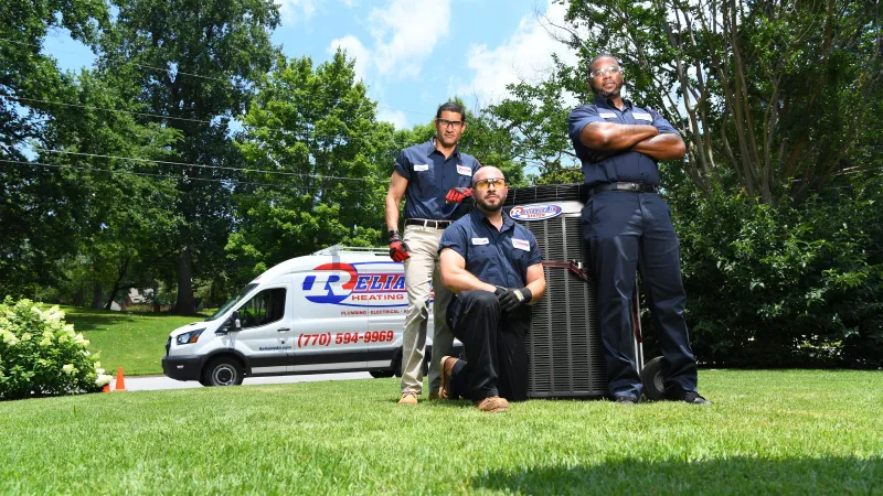 Three HVAC technicians posing with heating unit on green lawn beside company service van on sunny day.