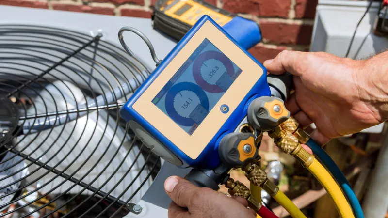 Technician using a digital manifold gauge set to measure pressure on an outdoor HVAC unit against a brick wall.