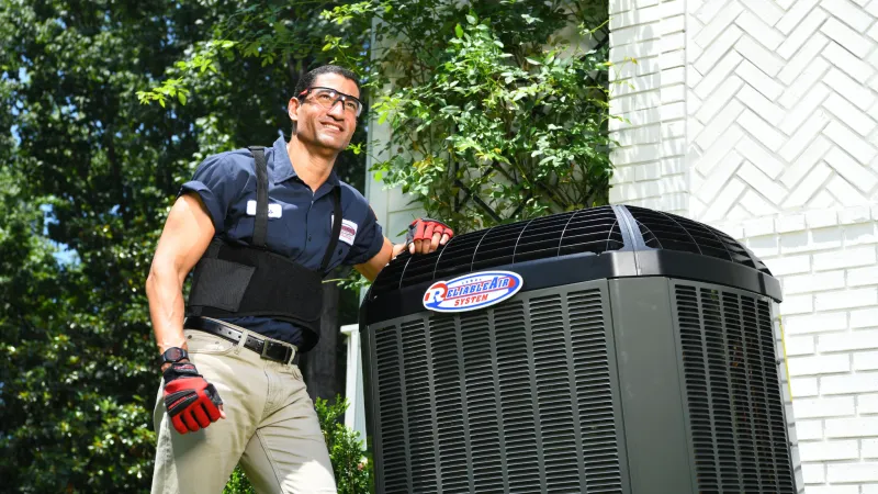 Technician wearing safety gear inspecting an outdoor air conditioning unit by a white brick house on a sunny day