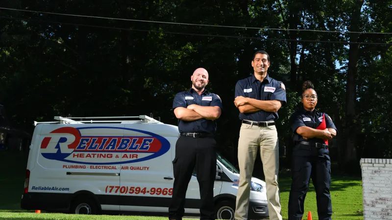 Three Reliable Heating & Air technicians pose confidently outdoors with service van in the background.
