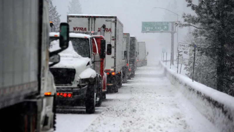 Line of semi trucks stuck on snowy highway during heavy winter storm with low visibility and snow-covered road.