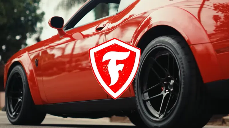 Close-up of a shiny red sports car with black wheels parked outdoors with a shield logo overlay.