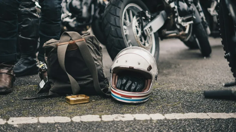 Vintage motorcycle helmet, leather gloves, backpack, and parked motorcycles on asphalt with riders nearby.