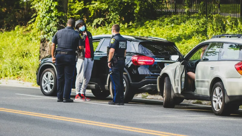 Two police officers talk to a man wearing a face mask beside parked cars on a roadside with greenery.