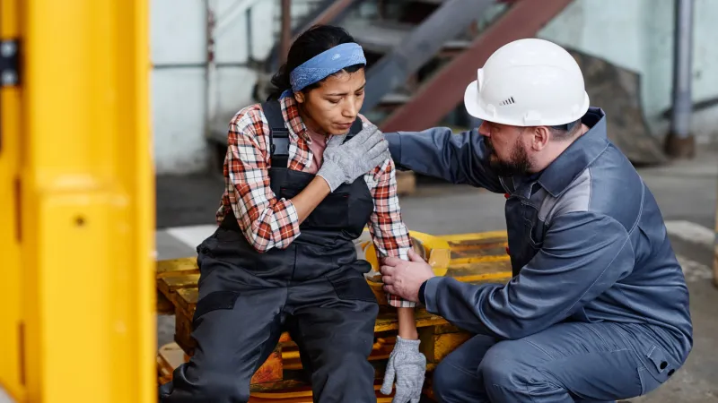 Male worker in white helmet comforting female worker sitting on pallet in industrial workplace with discomfort.