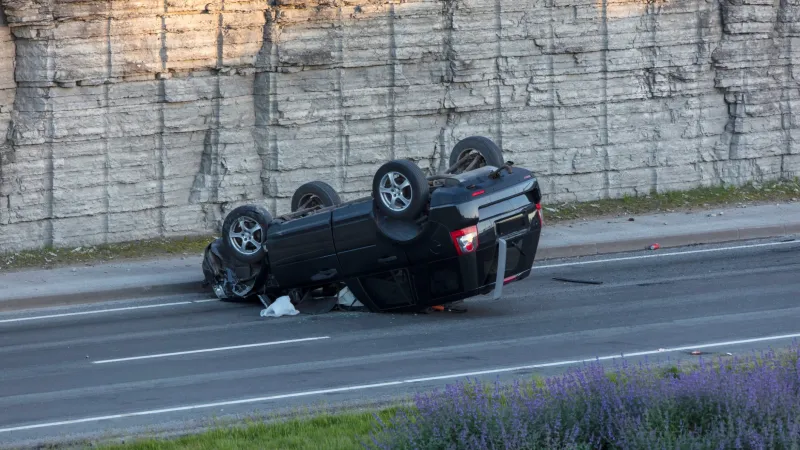 Black car flipped upside down on a highway near a rocky cliff after an accident.