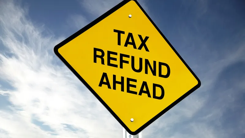 Yellow diamond road sign with Tax Refund Ahead text against a blue sky with clouds.
