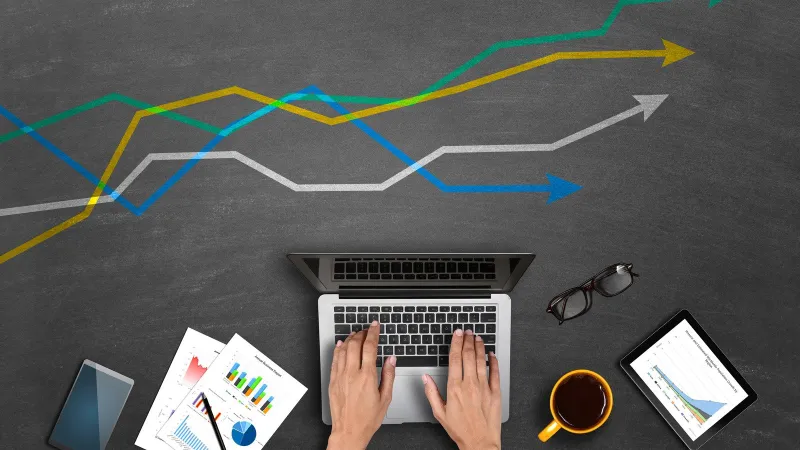 Hands typing on laptop with phone, papers, glasses, coffee, and tablet showing charts against chalkboard arrows