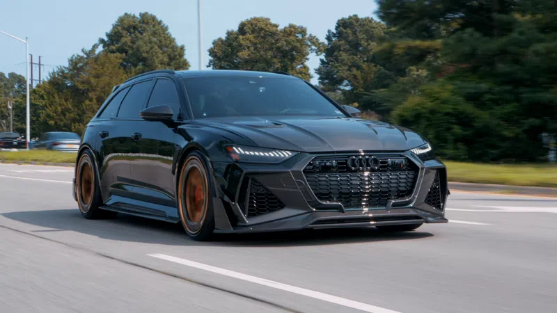 Black Audi RS6 Avant driving on a road with bronze rims and trees in the background under clear sky.
