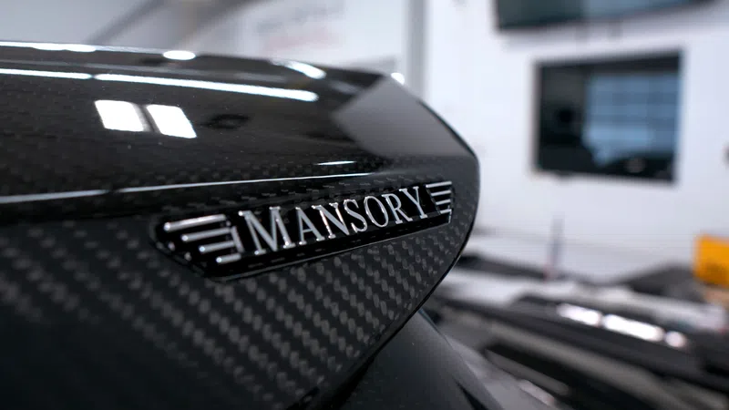 Close-up of Mansory car emblem on a glossy carbon fiber surface with blurred background indoors.
