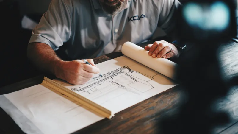Man in gray shirt sketching architectural plans with ruler on wooden table in focused work environment