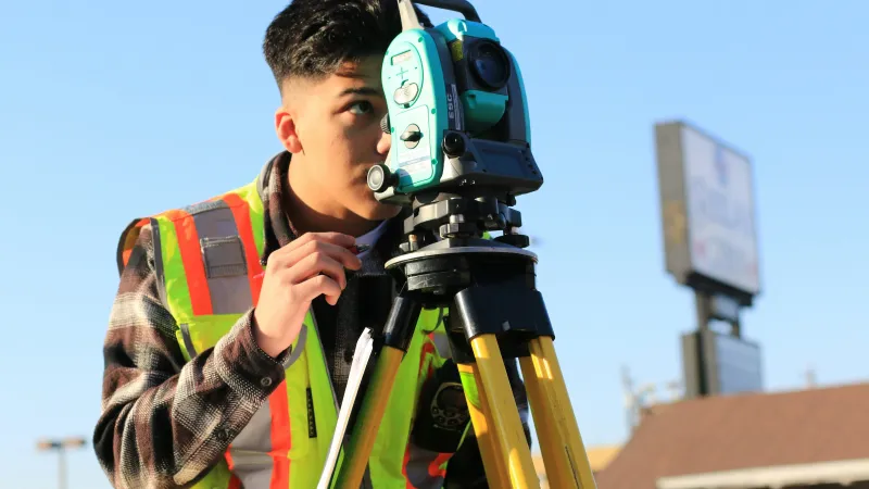 Surveyor in high-visibility vest using a theodolite tripod on a clear sunny day outdoors.