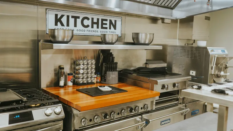 Clean commercial kitchen with stainless steel appliances, spices, cutting board, and a KITCHEN sign on the wall