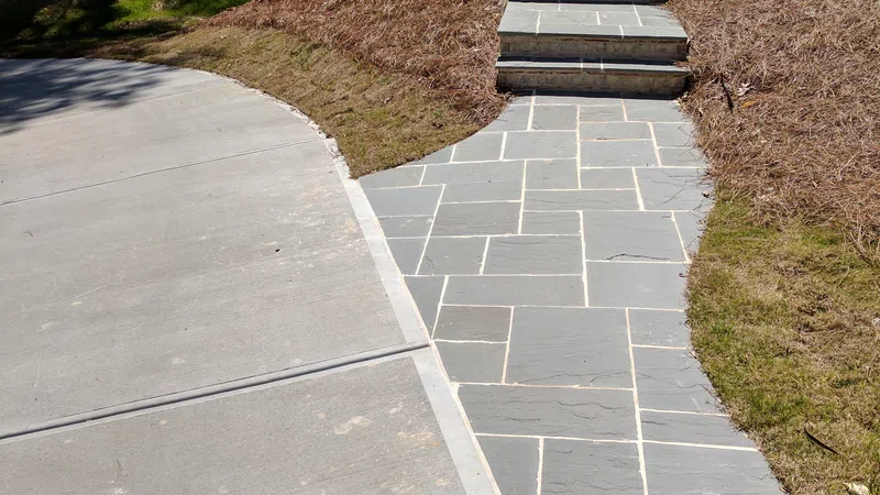 Stone pathway with stairs leading up to a house next to a concrete driveway in a landscaped yard.