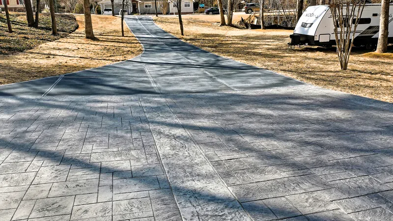 Long patterned concrete driveway leading to suburban home with RV and trees in a sunny yard.