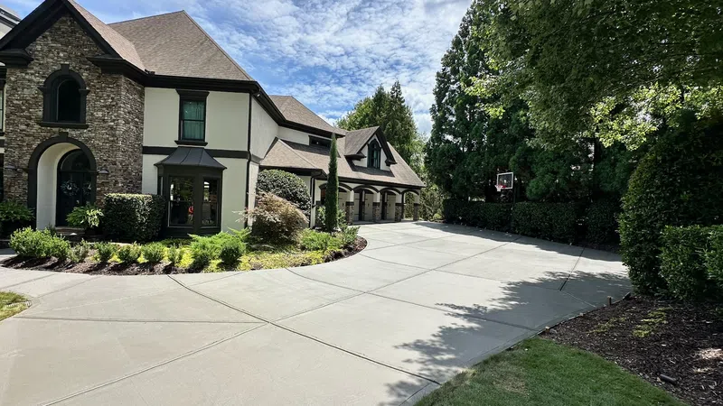 Spacious driveway leading to a large house with stone and stucco exterior surrounded by greenery under a partly cloudy sky